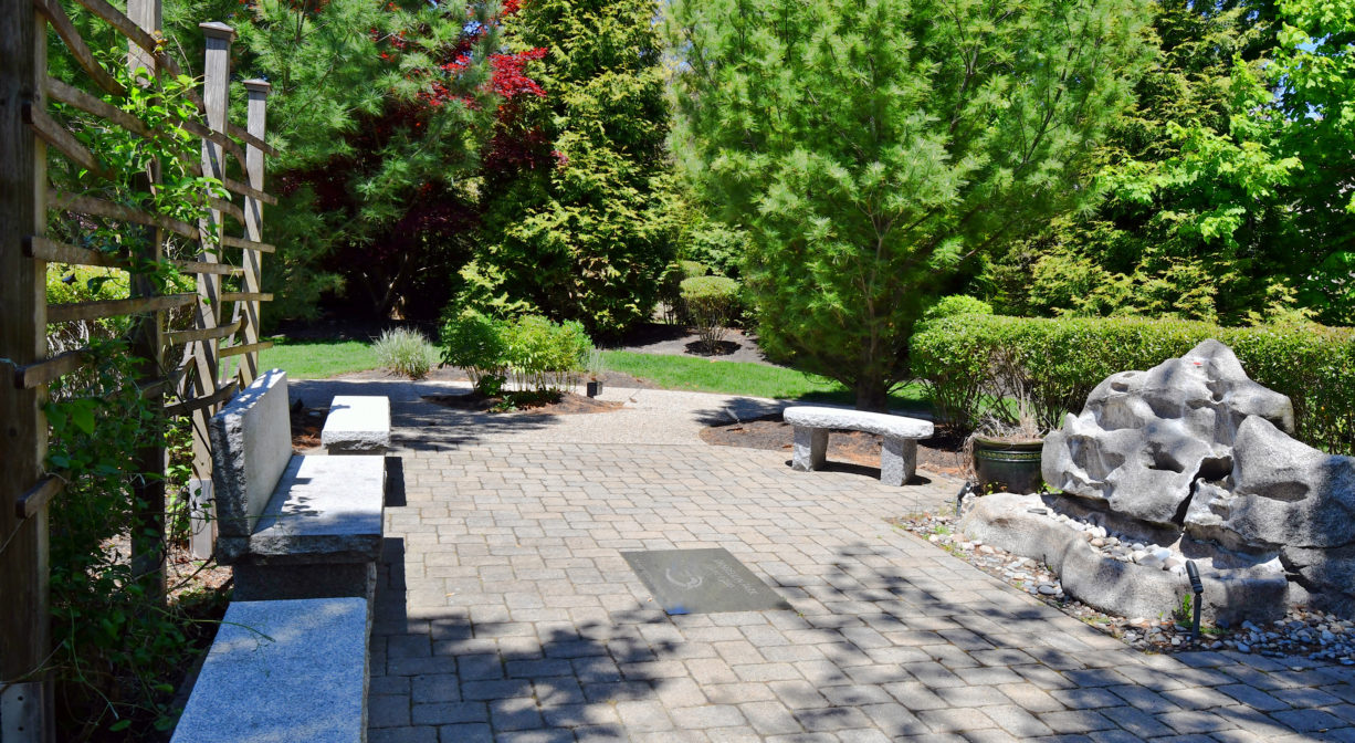 A photograph of a patio within a grassy park with a row of benches.