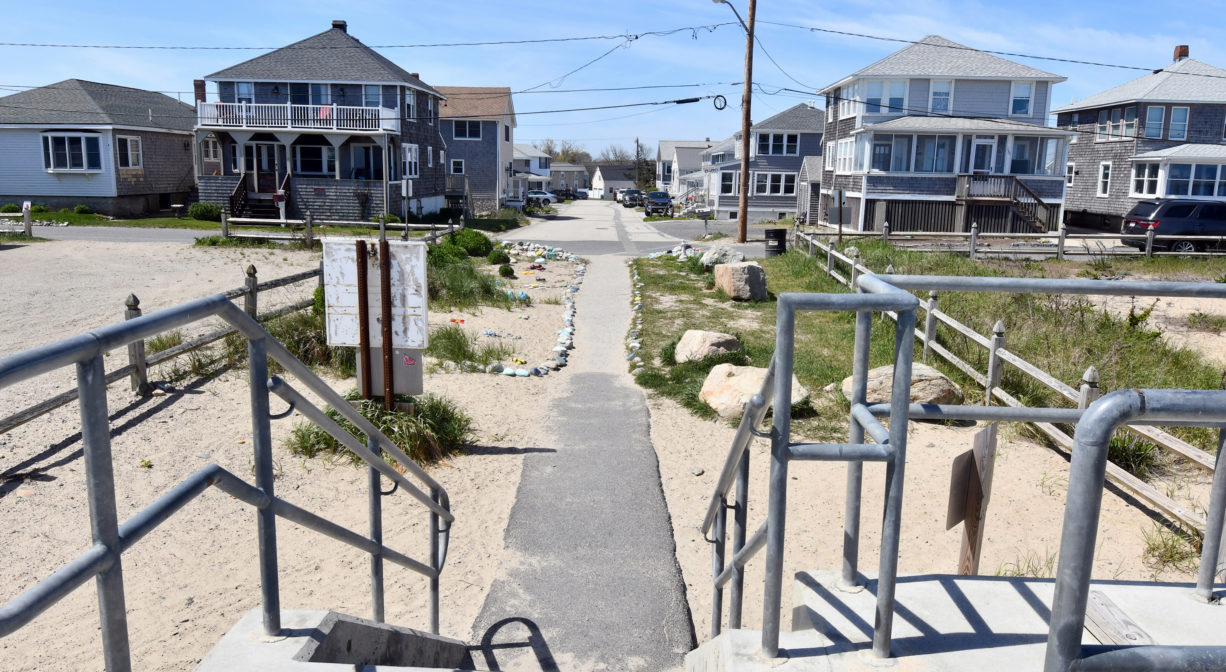 A photograph of a paved pathway extending from a seawall toward a residential area.
