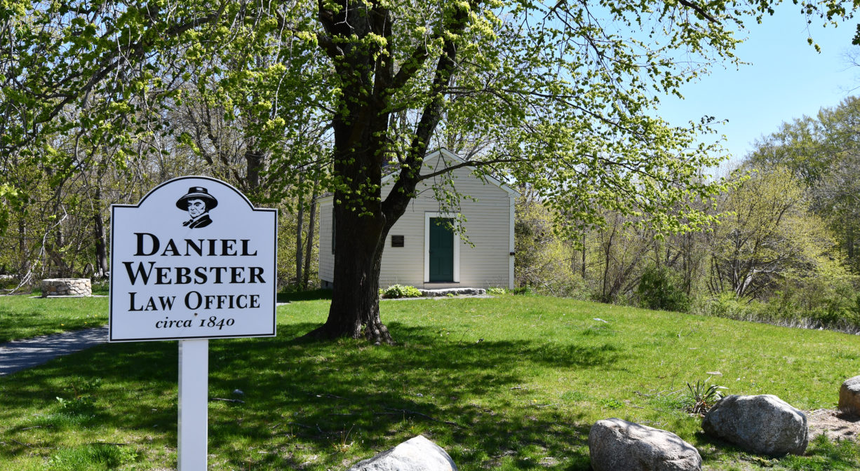 An interpretive sign with a small historic building in the background.