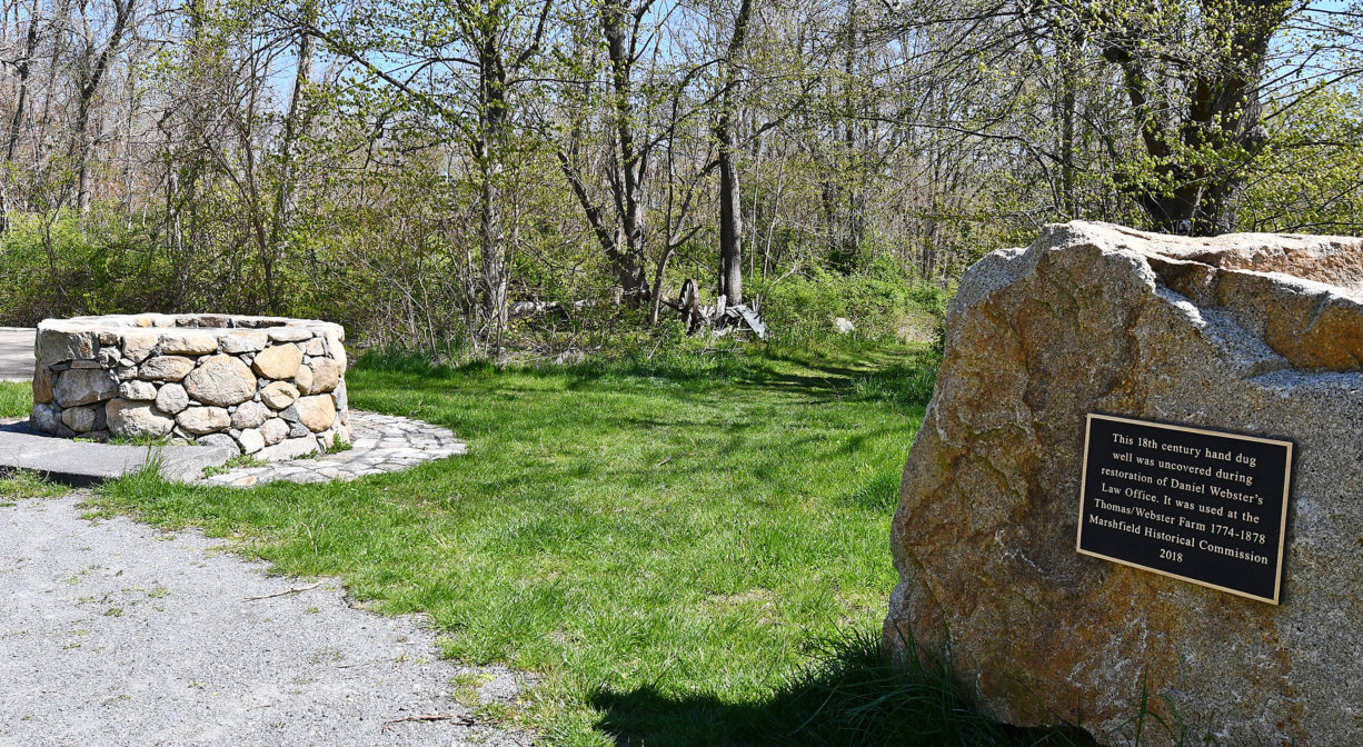 A photograph of a historic marker with grass, trees, and an old well.