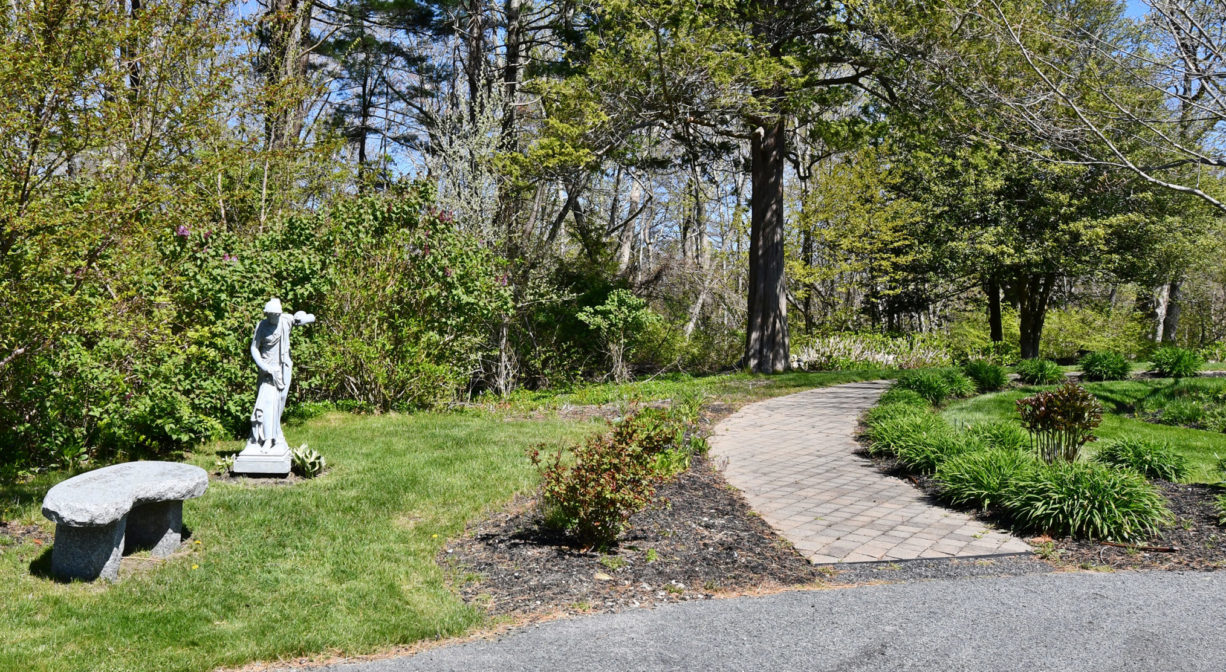 A photograph of a bench, a statue and a small garden with a walking path.