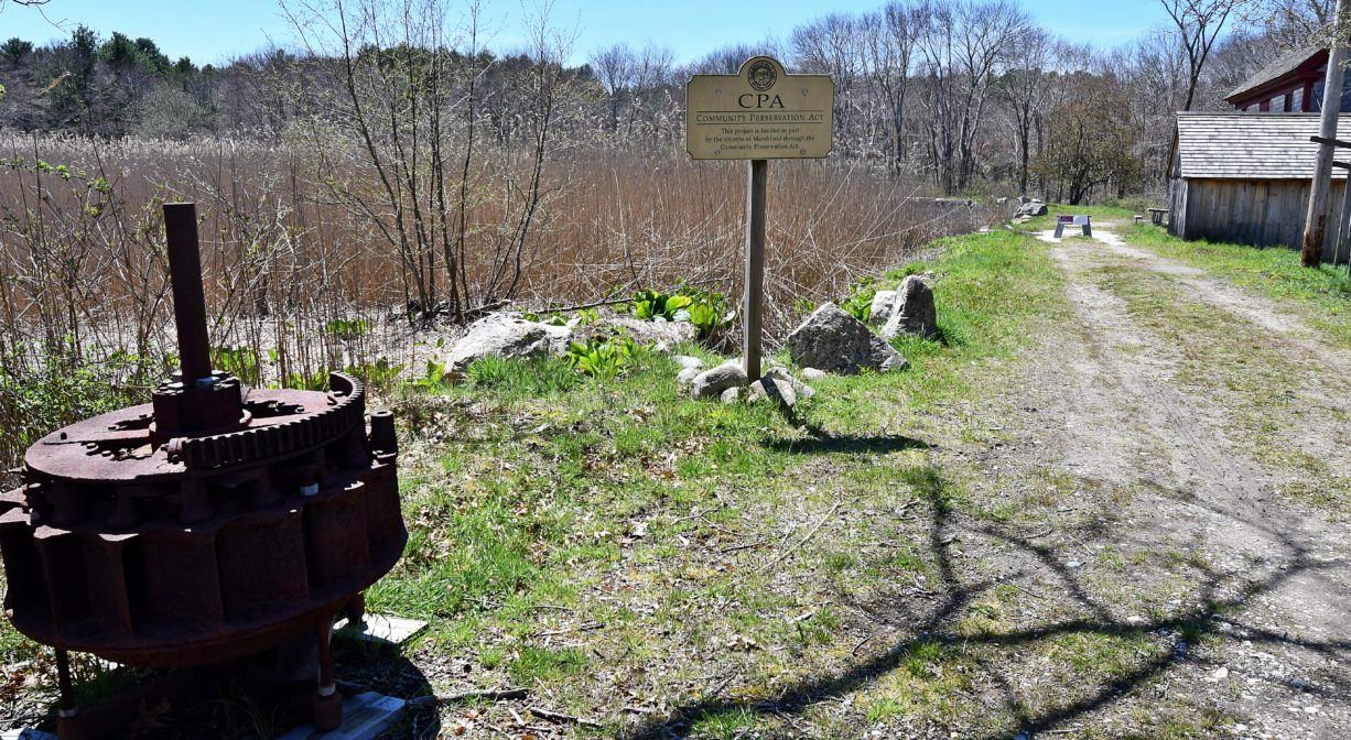 A photograph of an entrance road with a mill in the distance and a pond and some machine parts to one side.