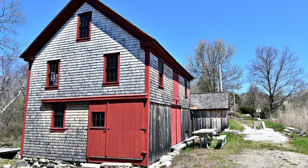 A photograph of a mill building beside a road with trees in the distance.