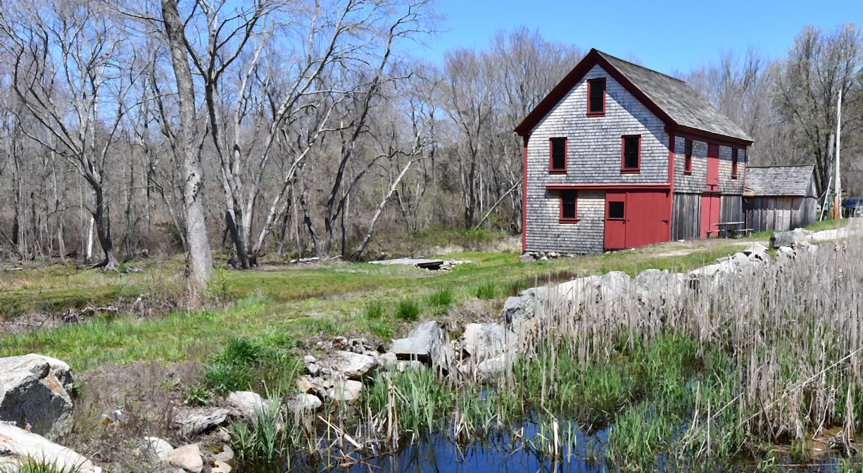 A photograph of a mill building and a mill stream, with grass and trees.