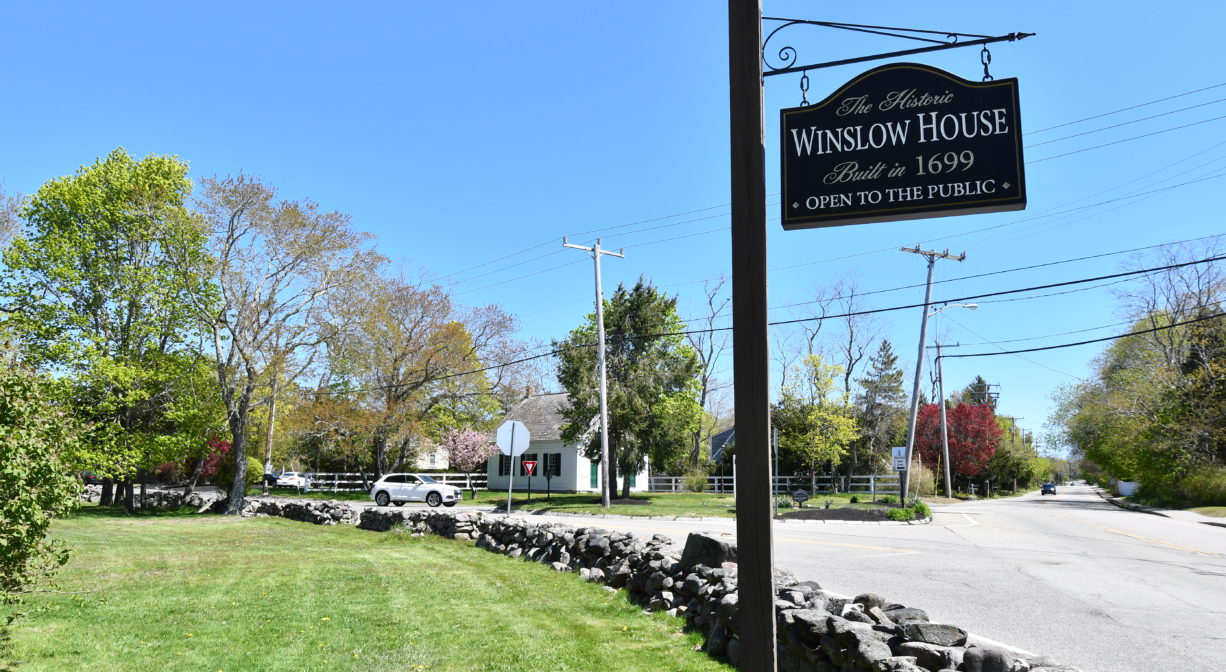 A photograph of a property sign and stone wall beside a roadway, with grass and trees.