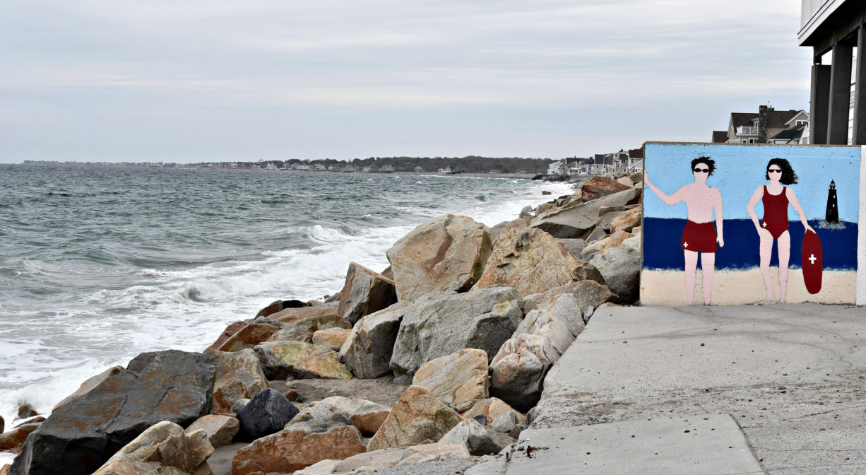 A photograph of a mural and some large rocks alongside the ocean.
