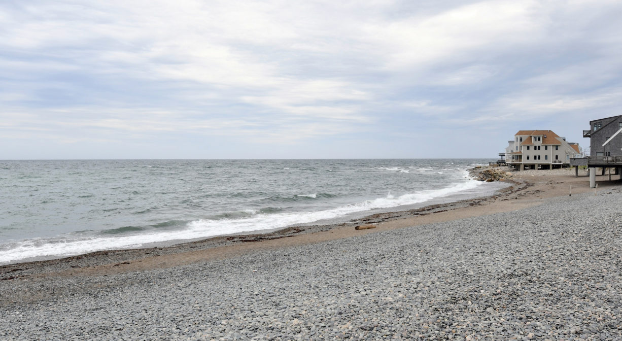 A stony beach with the ocean on one side and some houses in the distance.