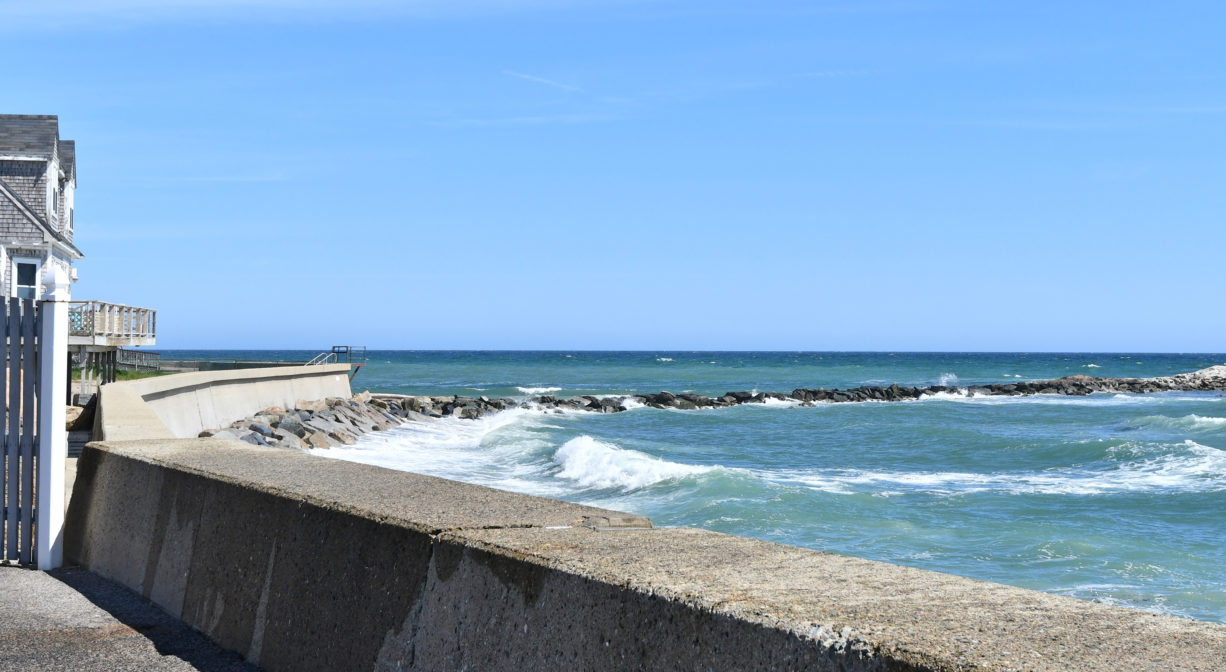 A photograph of a seawall with the ocean and a rocky outcropping in the background.