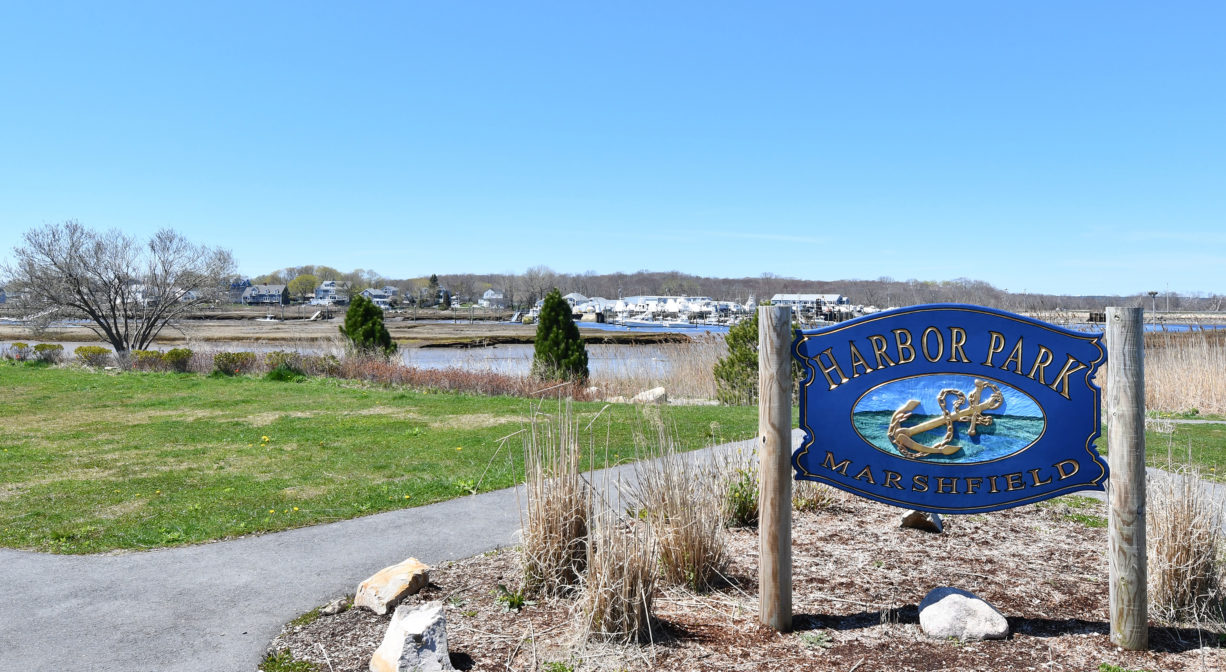 A photograph of a grassy park on the water with a property sign in the foreground and a paved trail.