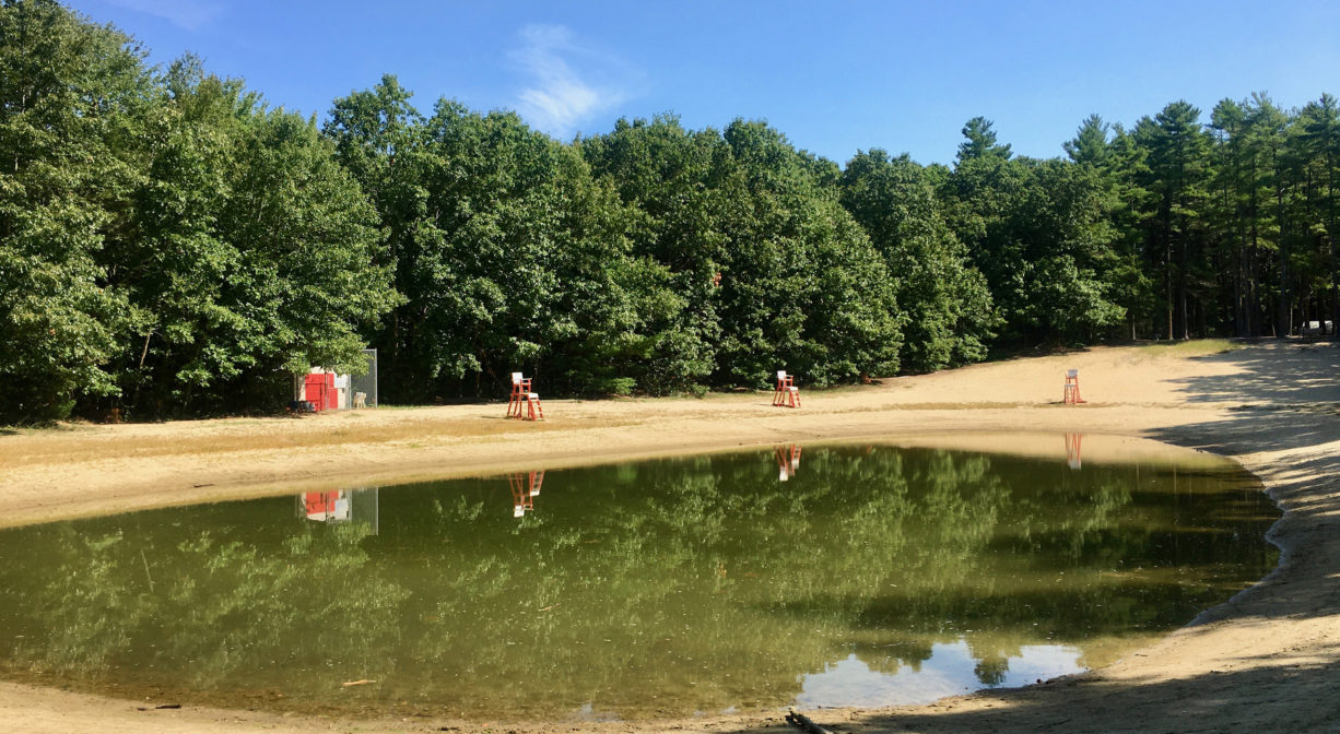 Photograph of manmade swimming pond surrounded with sand, with pine trees in the background.