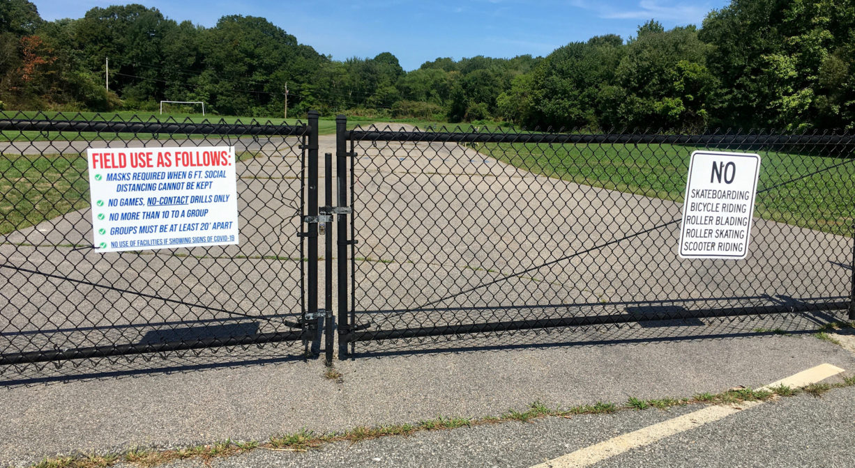 Photograph of a black metal entrance gate with a wide asphalt track in the background and blue skies.