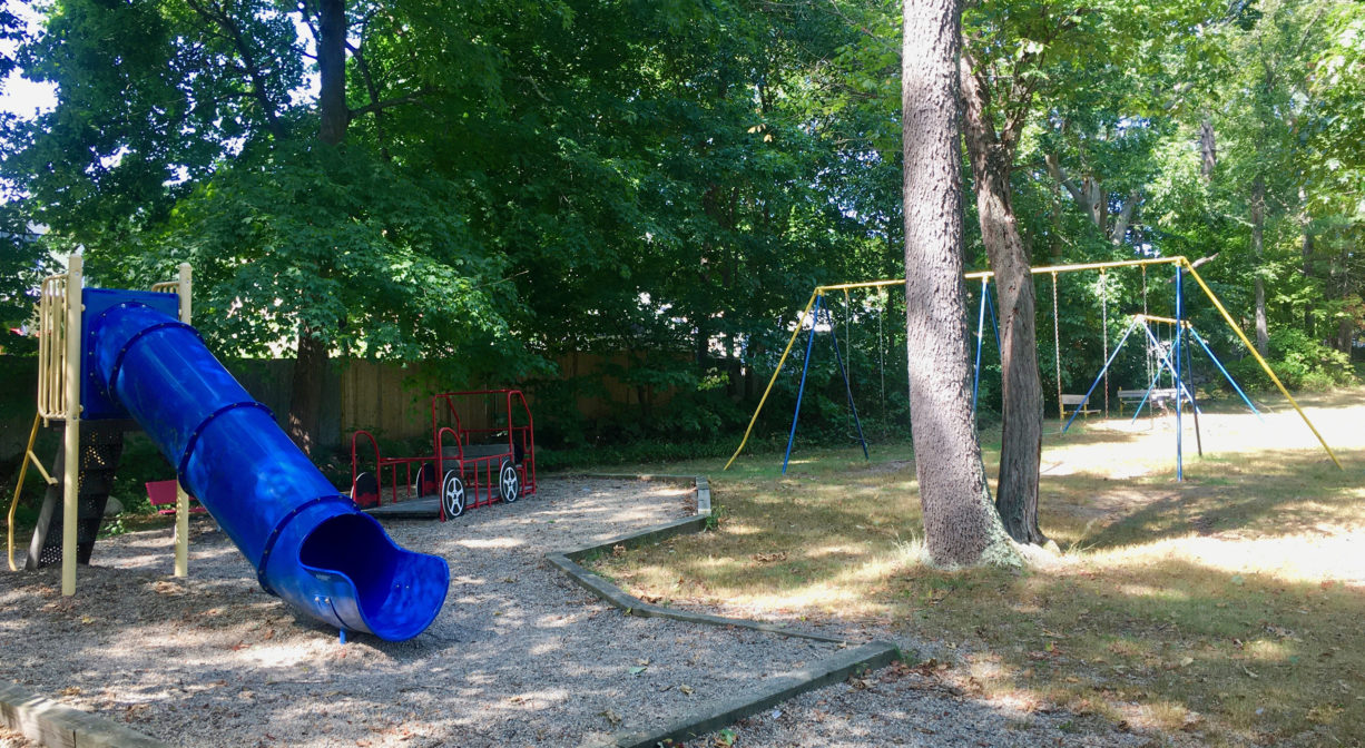 Photograph of playground equipment including swings and a slide with grass and trees.