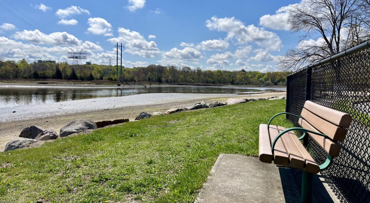 A photograph of a bench overlooking a grassy area, a sandy beach, and a river.