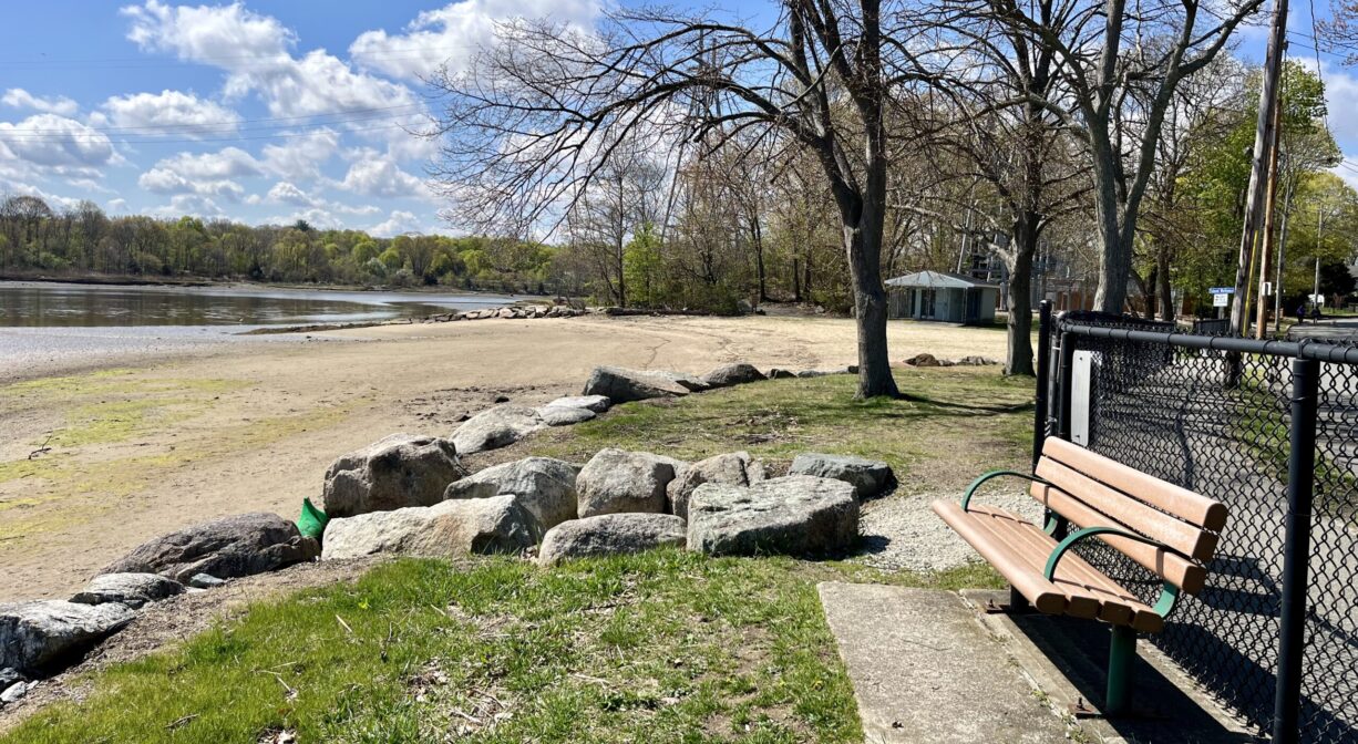 A photograph of a bench overlooking a grassy area, a sandy beach, and a river.