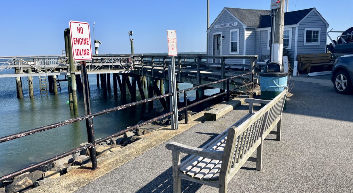 A photograph of a bench overlooking a pier on the ocean, with a building in the distance.