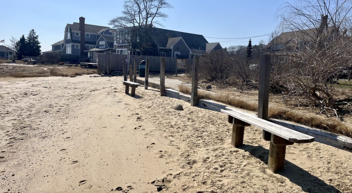 A photograph of two benches at the edge of a sandy beach, with houses in the distance.