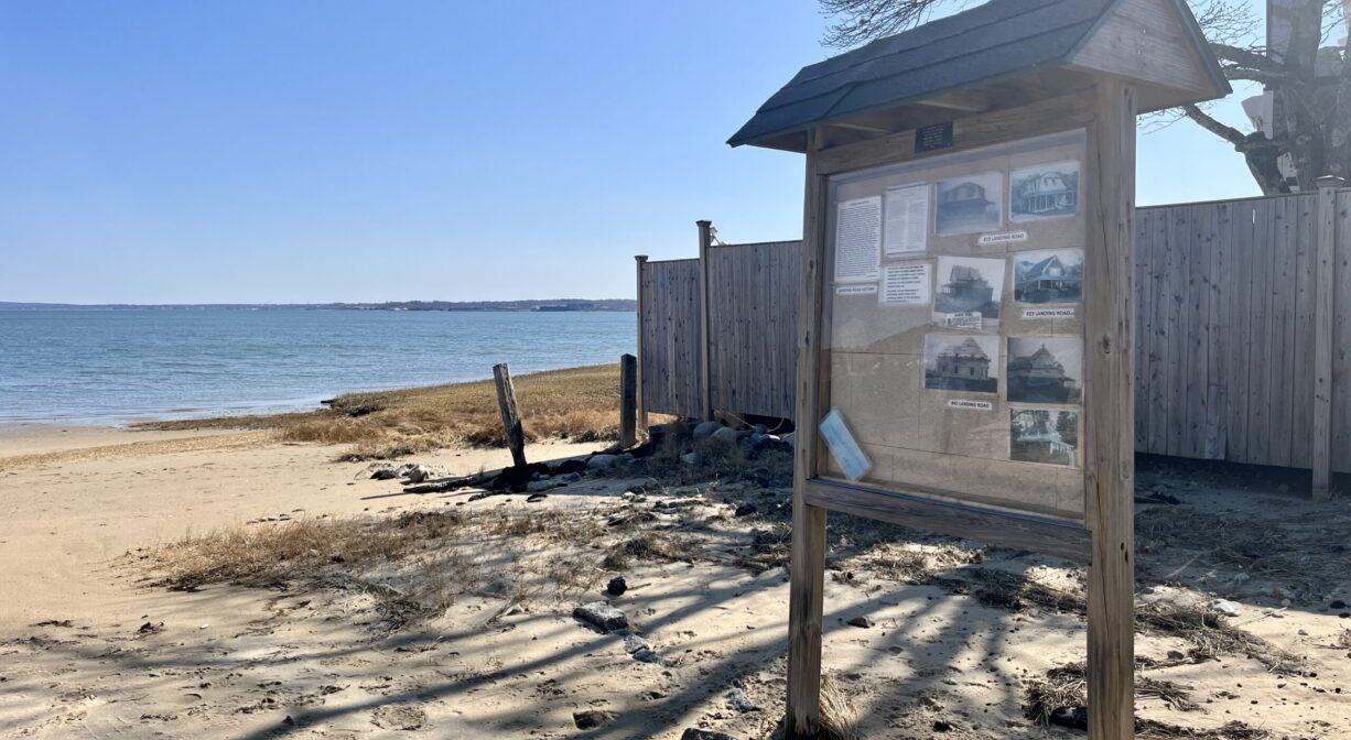 A photograph of an informational kiosk on a sandy beach with the ocean in the background.