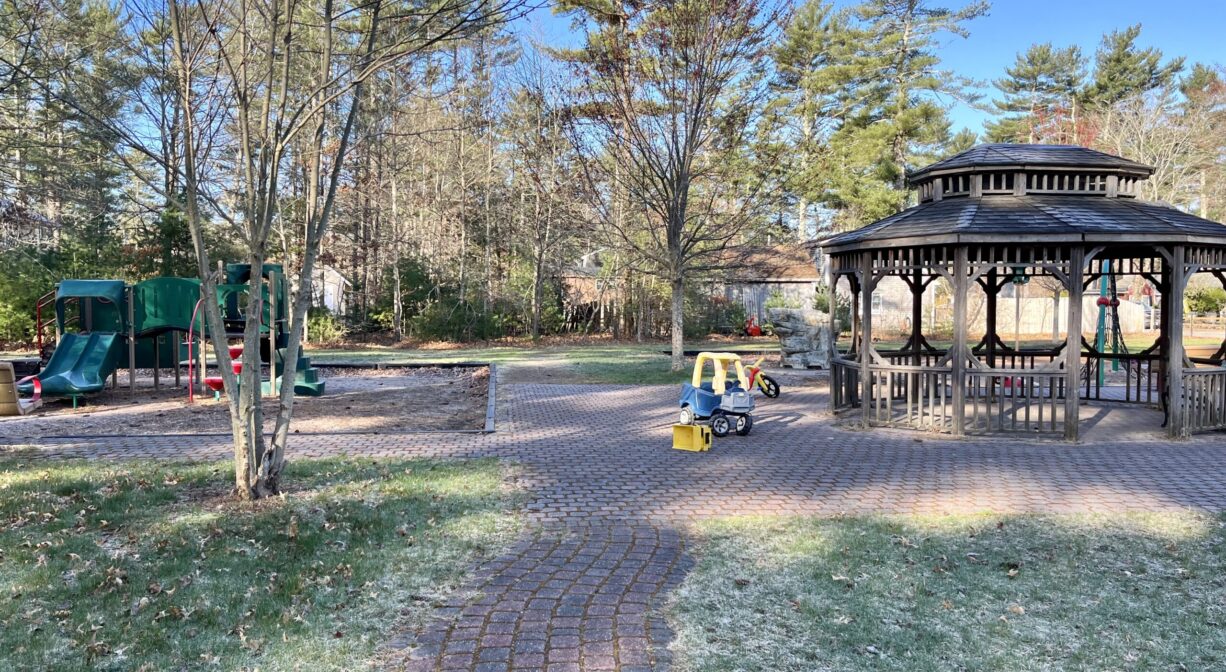 A photograph of a brick pathway into a playground, with a gazebo on one side and a play structure on the other.