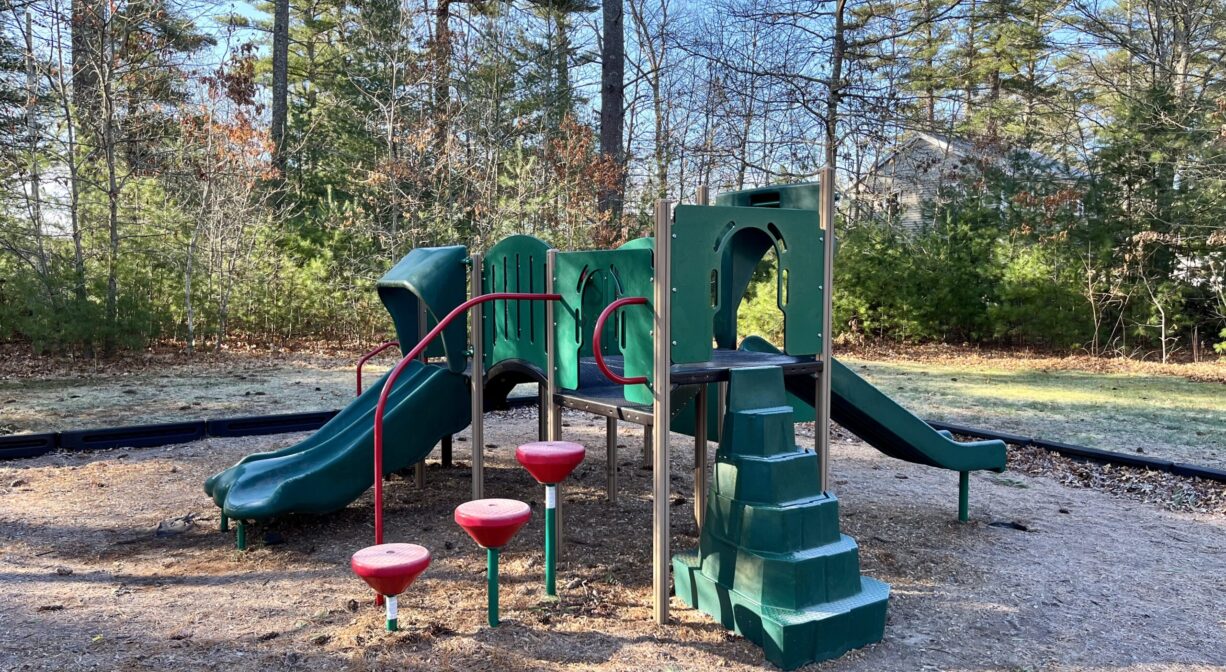 A photograph of a small green and red play structure for younger children with trees in the background.