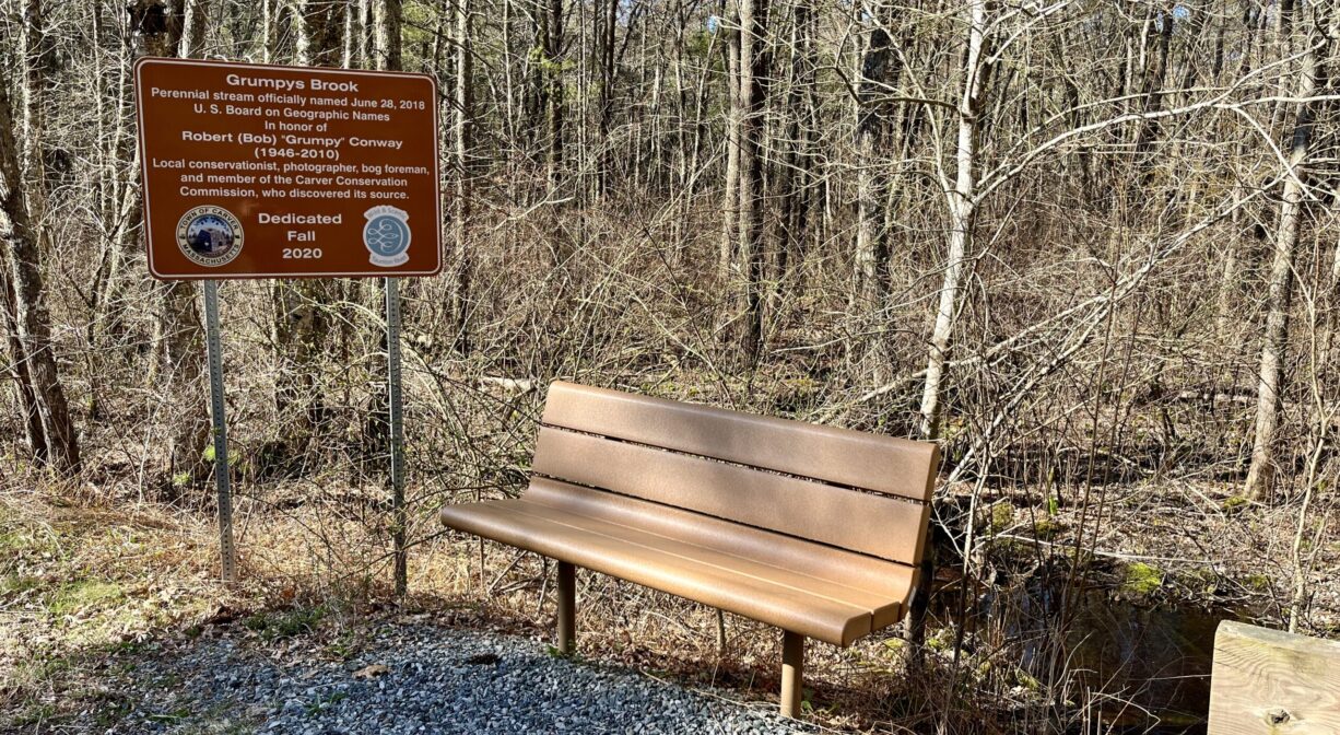 A photograph of a bench and a property sign beside a trail with a forest in the background.