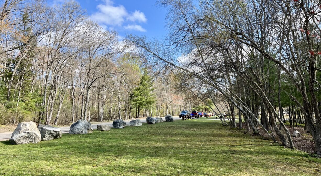 A photograph of a broad grassy area lined with large rocks, with trees to one side.