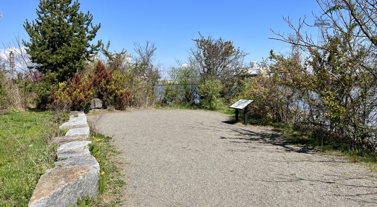A photograph of a wide trail and overlook, with trees and ornamental plantings, with a river in the distance.