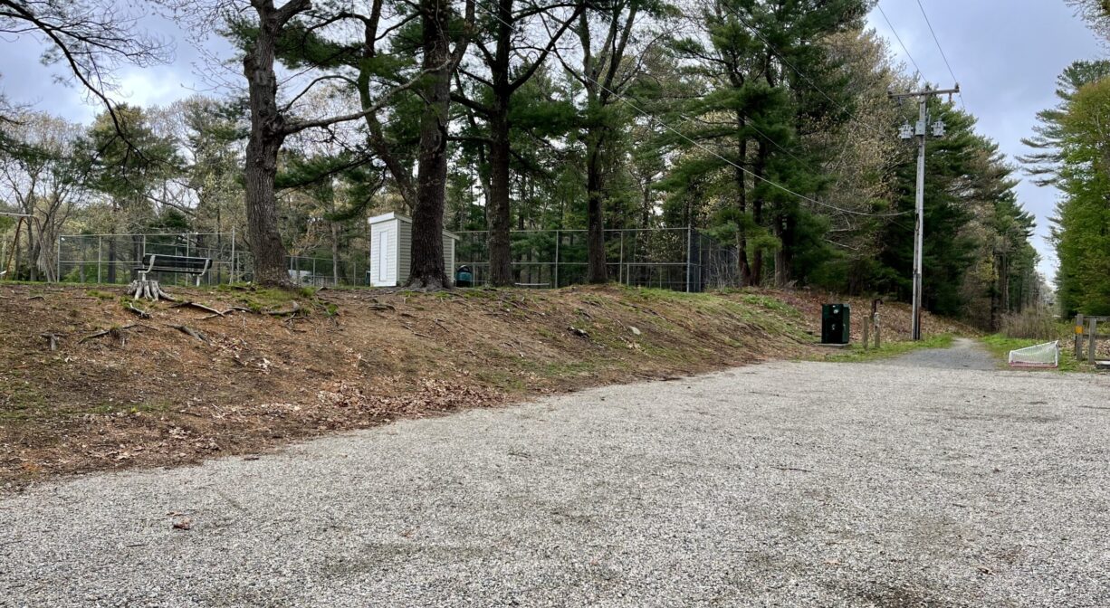 A photograph of a gravel parking area with woods in the background.