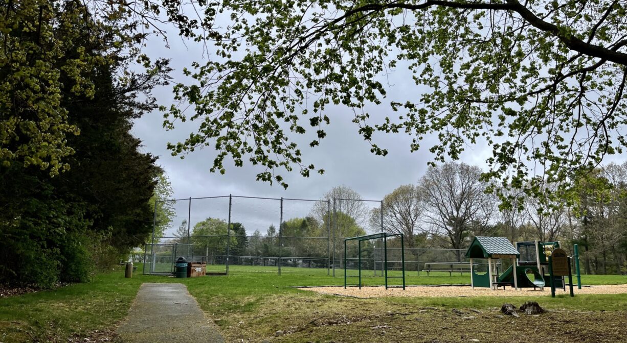 A photograph of a grassy park with a baseball field and small playground, with trees to one side.