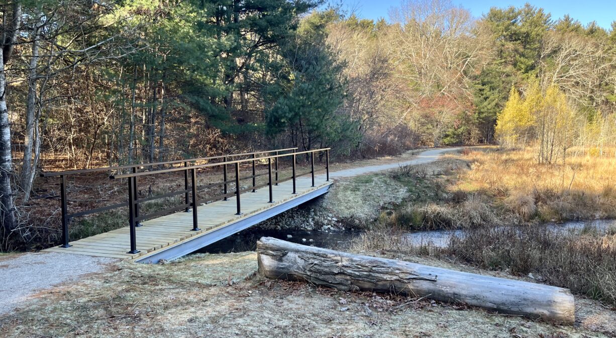 A photograph of a bridge and trail over a stream with a log in the foreground.